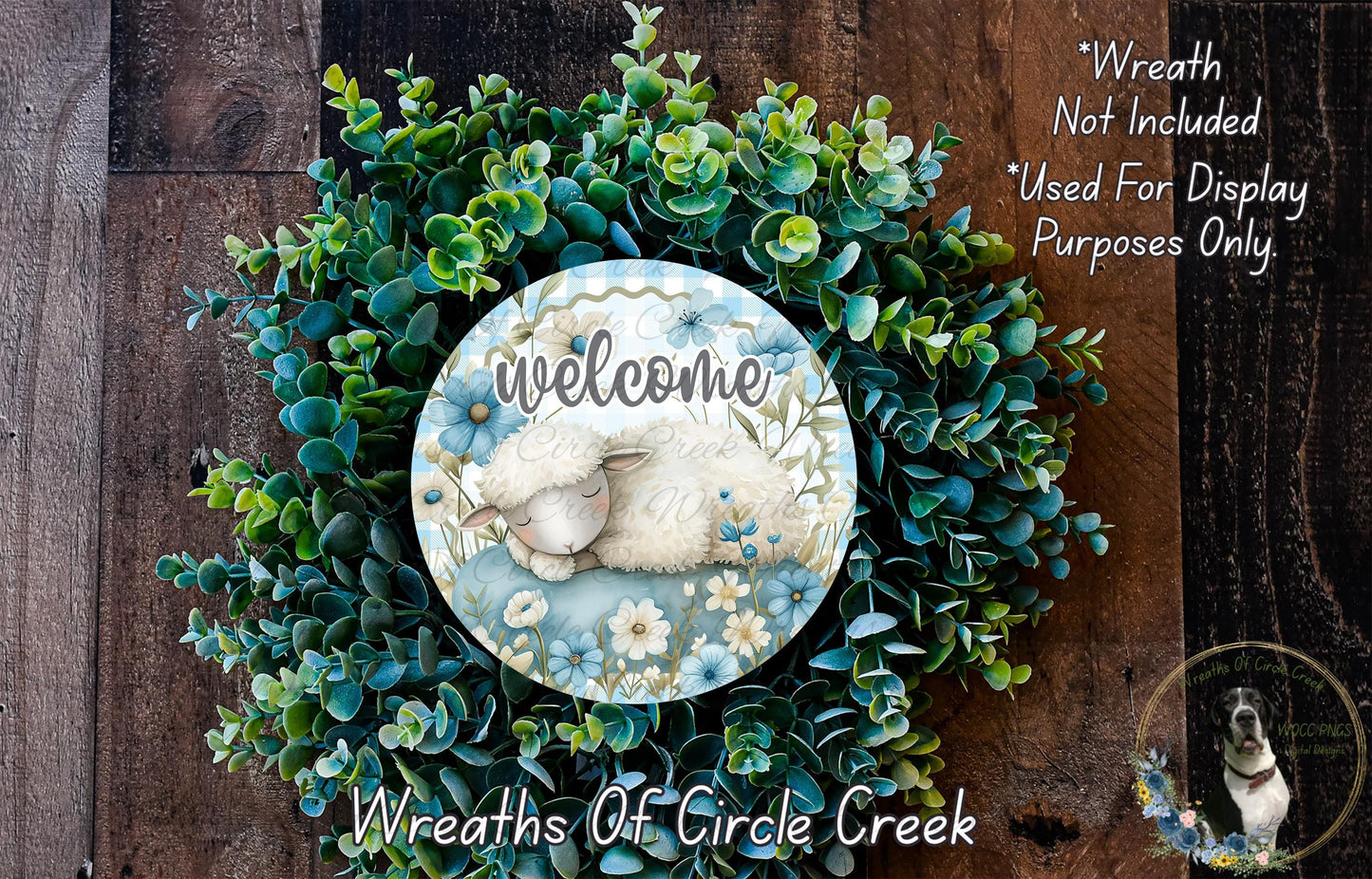a wreath with a sleeping lamb at the center, surrounded by various green plants and flowers. The wreath is displayed on a wooden surface, and there is a text overlay that reads "Welcome Wreath Not Included * Used For Display Purposes Only".