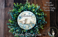a wreath with a sleeping lamb at the center, surrounded by various green plants and flowers. The wreath is displayed on a wooden surface, and there is a text overlay that reads &quot;Welcome Wreath Not Included * Used For Display Purposes Only&quot;.