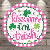 A circular sign with the text "kiss me, I'm Irish" surrounded by shamrocks and a pink and white checkered border.