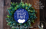 A wreath made of green leaves and a blue circular ornament with the text "Gloria to the Newborn King" and "De Creek" written on it, placed on a wooden surface.