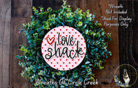 a wreath made of green leaves and a pink circular sign with the text "Love Shack" written on it.