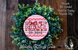 A wreath made of green leaves and a pink and white sign with the text "LOVE like Jesus" is displayed on a wooden surface.