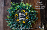 a wreath made of various plants, including sunflowers and greenery, with the text "Home Sweet Home" written on it. The wreath is displayed on a wooden surface, and there is a small dog in the bottom right corner of the image.