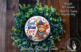 a wreath with a cow wearing a patriotic ribbon and surrounded by greenery. The wreath has a patriotic ribbon and the text "Land that I Love" on it. The wreath is displayed on a wooden surface, and there is a small dog in the bottom right corner of the image.