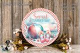 a decorative round sign with a beach-themed design, surrounded by flowers and placed on a wooden surface.