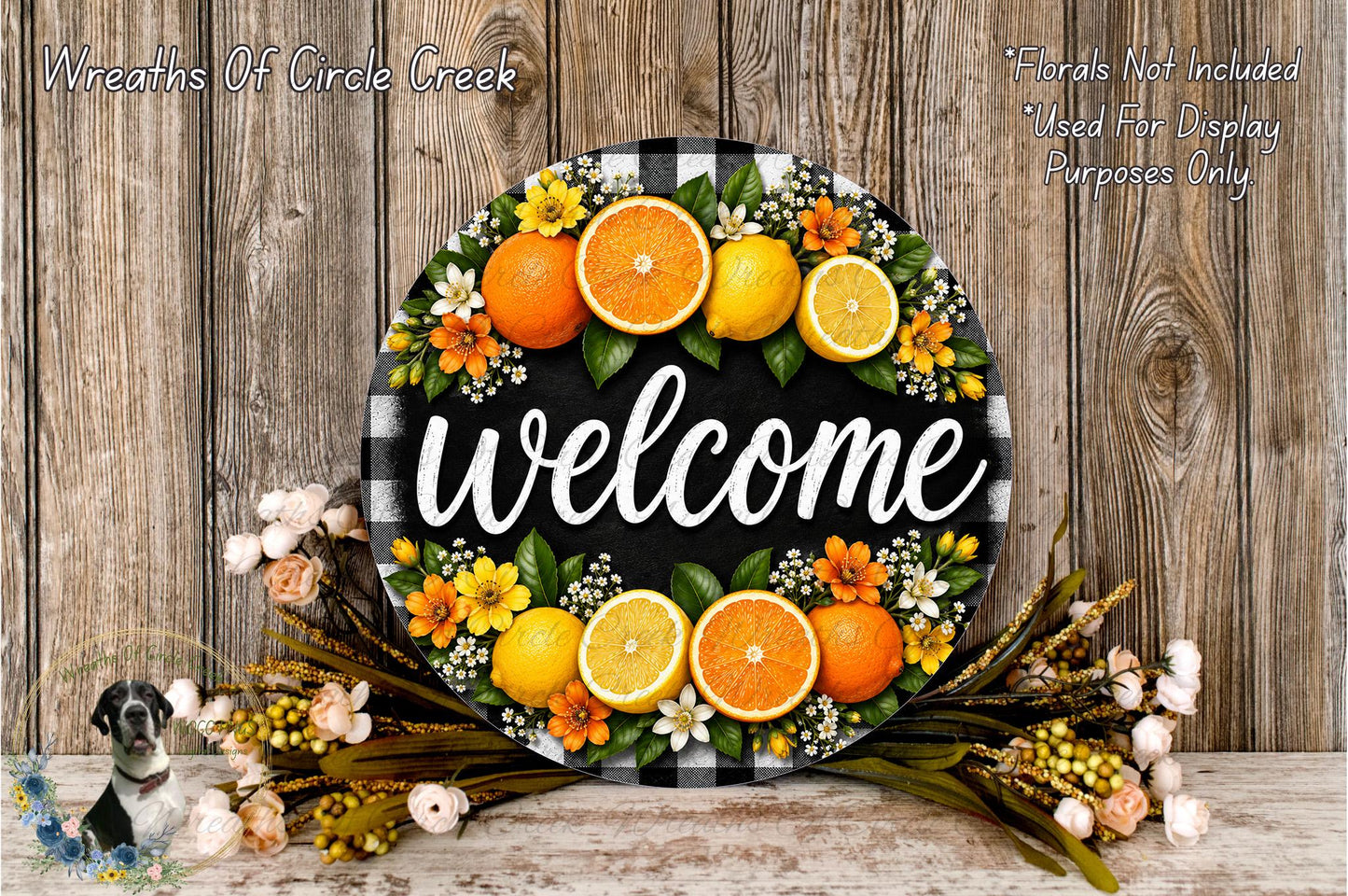 A decorative welcome sign made of a circular wooden plaque with a black and white checkered pattern, surrounded by a wreath of orange and yellow flowers, and a bunch of oranges.