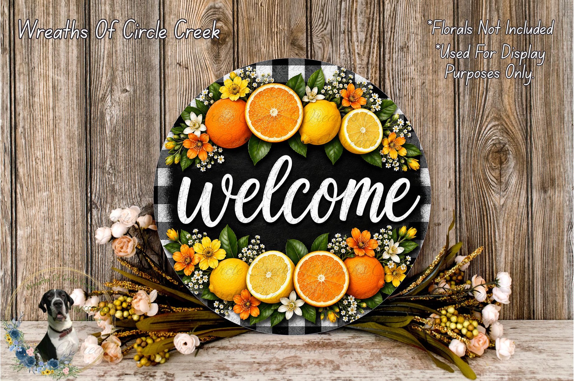A decorative welcome sign made of a circular wooden plaque with a black and white checkered pattern, surrounded by a wreath of orange and yellow flowers, and a bunch of oranges.