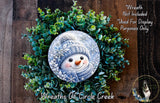 A wreath made of green plants and a snowman decoration, with a caption stating that the wreath is not included in the display and is used for display purposes only.