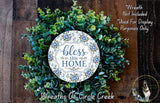 A wreath made of various green plants and flowers, with a white circular sign that says "bless this home" in blue and green lettering.