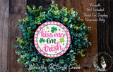 a wreath made of green leaves and a pink and white circular sign with the text "Kiss me Irish" on it. The sign is surrounded by a lush wreath, creating a festive and decorative display.