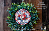 a wreath made of various green plants and flowers, with a red and white plaid circular sign that says "welcome" in the center. The wreath is displayed on a wooden surface, and there is a small dog in the bottom right corner of the image.