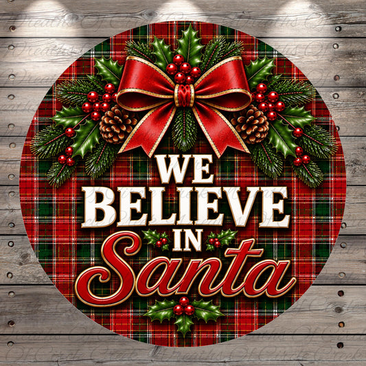 A circular Christmas-themed sign with a plaid pattern, featuring a red bow, pine cones, berries, and holly leaves. The text "WE BELIEVE IN Santa" is displayed prominently in the center of the sign.