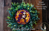 a wreath with a colorful design, including a woman's face with a skull-like appearance, surrounded by green foliage.