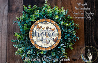 a wreath made of various green plants and a decorative plate with a message. The wreath is displayed on a wooden surface, and the plate has a message that reads "Bless this home and all who enter".