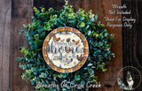 a wreath made of various green plants and a decorative plate with a message. The wreath is displayed on a wooden surface, and the plate has a message that reads "Bless this home and all who enter".