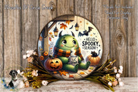 A whimsical illustration of a green monster with horns and a scarf, sitting on a wooden surface surrounded by autumn decorations such as pumpkins, leaves, and flowers. The monster appears to be holding a mug with the text "Hello Spooky Season!" written on it.