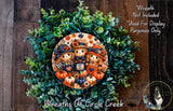a wreath with a central design of three scarecrow characters surrounded by various fall-themed elements such as pumpkins, leaves, and flowers. The wreath is set against a backdrop of green foliage and a wooden wall.