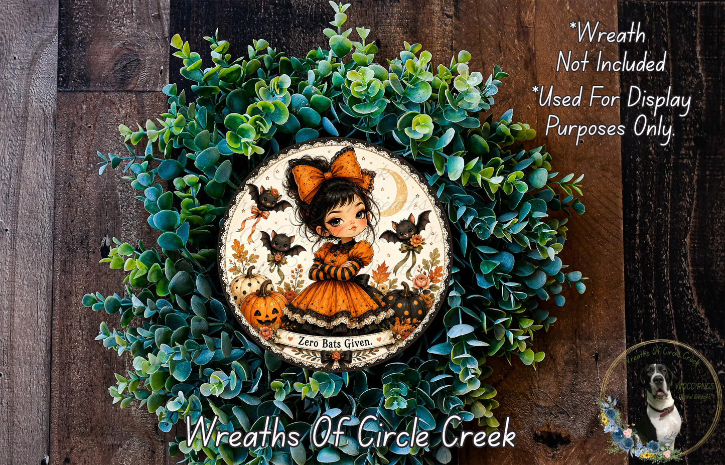 a wreath with a girl wearing a witch's hat and a pumpkin, surrounded by green foliage. The wreath is displayed on a wooden surface, and there is a small dog in the bottom right corner of the image.