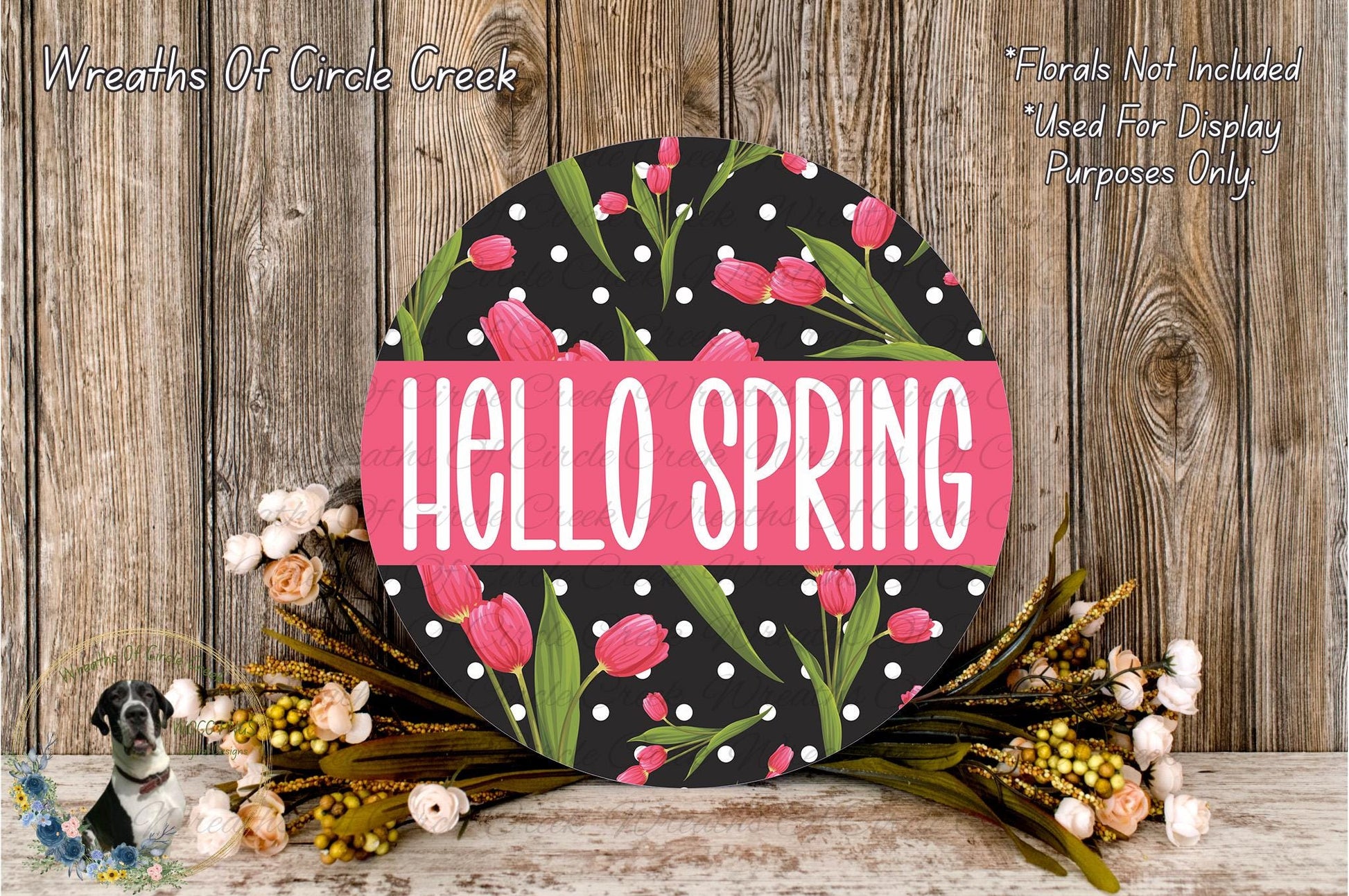 a round sign with the text "Hello Spring" in white letters, surrounded by a floral arrangement of pink tulips and green leaves on a wooden surface.