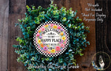 a wreath made of various green plants and flowers, with a sign that reads "Welcome to my happy place don't ruin it". The wreath is displayed on a wooden surface, and there is a small dog in the bottom right corner of the image.