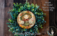 a wreath with a scarecrow design, surrounded by green foliage. The wreath is placed on a wooden surface, and there is a small dog visible in the bottom right corner of the image.