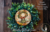 a wreath with a scarecrow design, surrounded by green foliage. The wreath is placed on a wooden surface, and there is a small dog visible in the bottom right corner of the image.