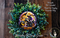 A Halloween-themed wreath made of green foliage and featuring a central image of a witch riding a motorcycle against a full moon backdrop.