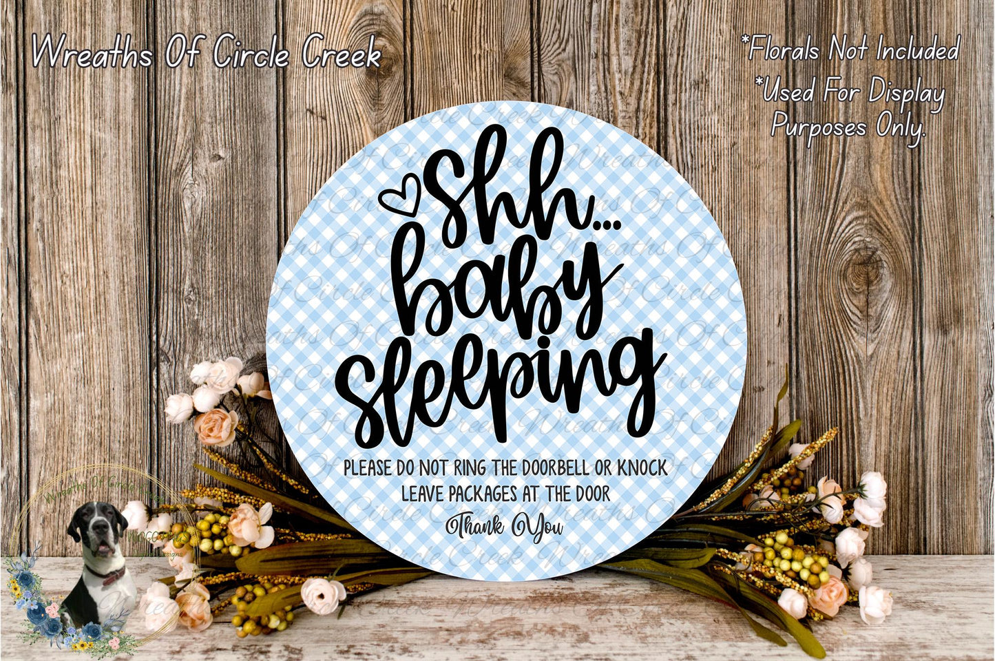 a round sign with a blue and white checkered pattern, surrounded by a floral arrangement on a wooden surface. The sign displays the text "Shh... baby sleeping" in a playful, handwritten style.