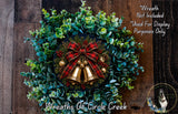 A festive Christmas wreath made of evergreen branches, adorned with a red and black plaid bow, bells, and berries. The wreath is displayed on a wooden wall, and there is a small dog in the bottom right corner of the image.