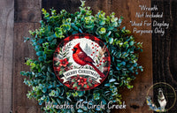 A Christmas wreath made of green foliage and red berries, with a red cardinal bird perched on a branch. The wreath has a "Merry Christmas" banner and is displayed on a wooden surface.