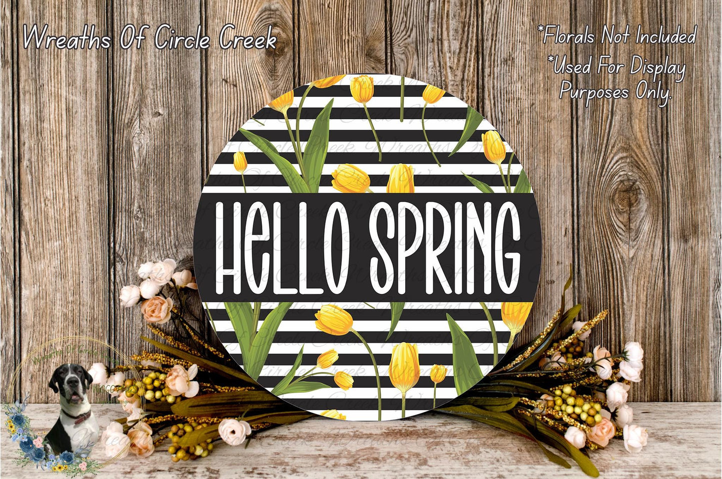 a round sign with the text "Hello Spring" in bold, surrounded by a wreath of yellow flowers and green leaves. The sign is placed on a wooden surface, and there is a small dog in the background.