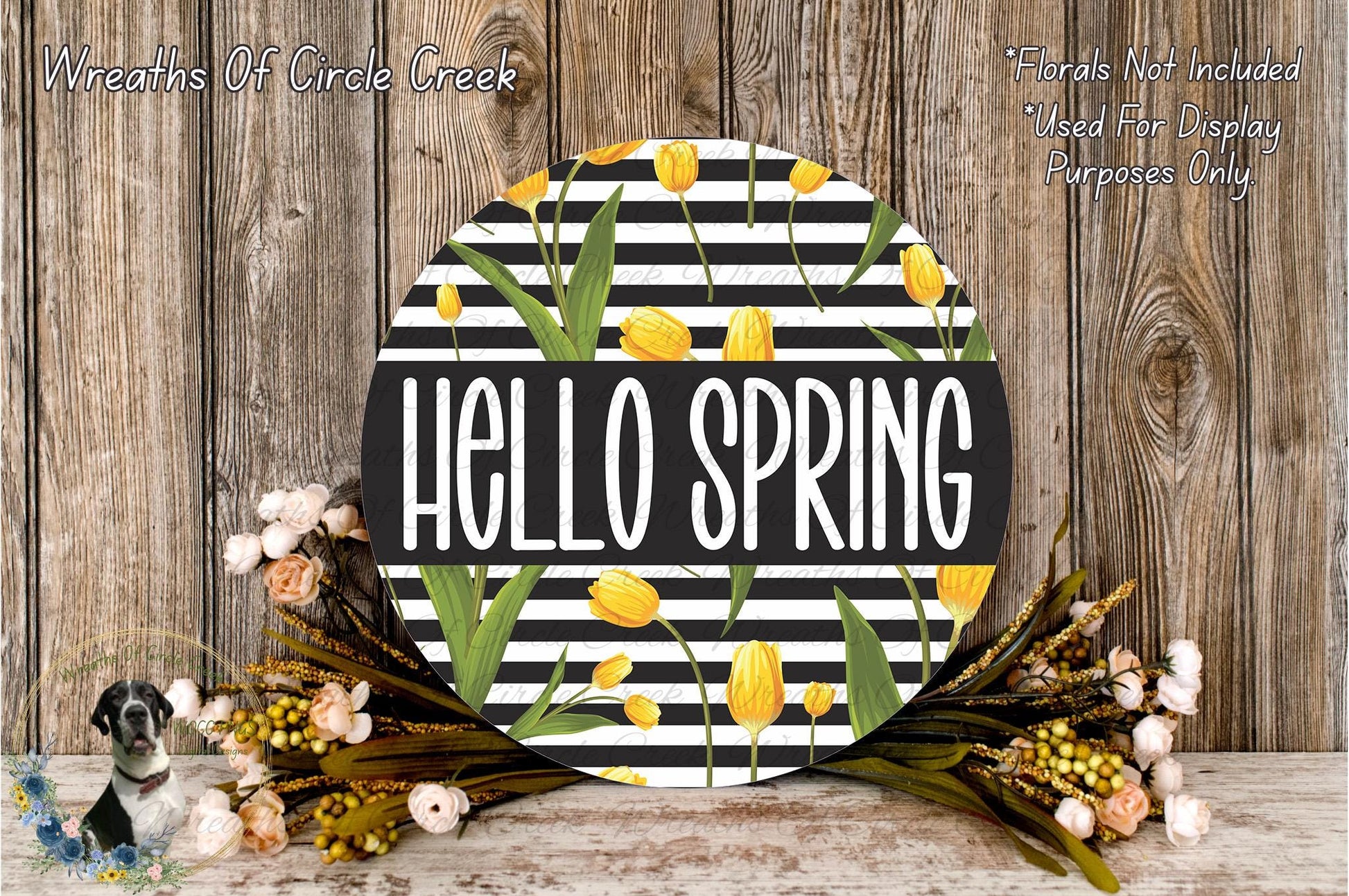 a round sign with the text "Hello Spring" in bold, surrounded by a wreath of yellow flowers and green leaves. The sign is placed on a wooden surface, and there is a small dog in the background.