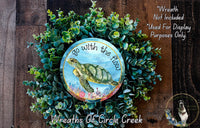 A wreath made of green plants and a circular design featuring a turtle, with the text "Go with the flow" written on it.