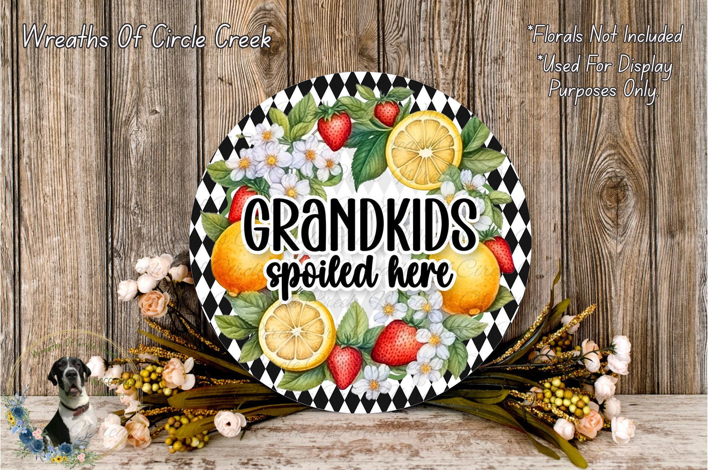 A decorative wreath with a black and white checkered border, featuring a variety of fruits such as oranges, strawberries, and lemons, along with a text that reads "GRANDKIDS spoiled here". The wreath is placed on a wooden surface, surrounded by flowers and greenery.
