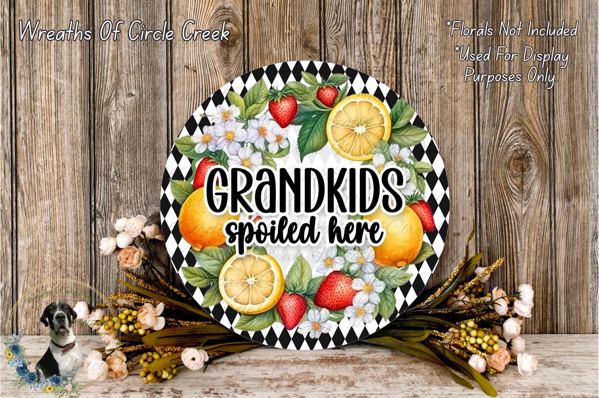 A decorative wreath with a black and white checkered border, featuring a variety of fruits such as oranges, strawberries, and lemons, along with a text that reads "GRANDKIDS spoiled here". The wreath is placed on a wooden surface, surrounded by flowers and greenery.