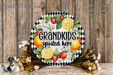 A decorative wreath with a black and white checkered border, featuring a variety of fruits such as oranges, strawberries, and lemons, along with a text that reads "GRANDKIDS spoiled here". The wreath is placed on a wooden surface, surrounded by flowers and greenery.