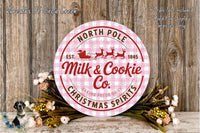 a round sign with text and a graphic of a Santa Claus sleigh, surrounded by a floral arrangement on a wooden surface.