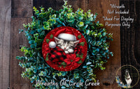 A wreath made of green and red foliage, with a sleeping kitten wearing a Santa hat and a red and black plaid blanket, surrounded by a red and white poinsettia flower.