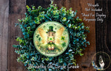 A wreath featuring a fairy with a green hat and a green shamrock, surrounded by a lush green plant. The fairy appears to be holding a book, and the wreath is described as a "Fairy Blessings" wreath.