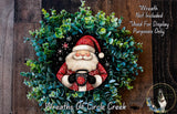 A wreath featuring a Santa Claus figure holding a cup of coffee, surrounded by greenery.
