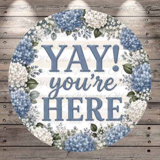A circular sign with the text "YAY you're here" surrounded by a floral border of blue hydrangeas and white flowers.