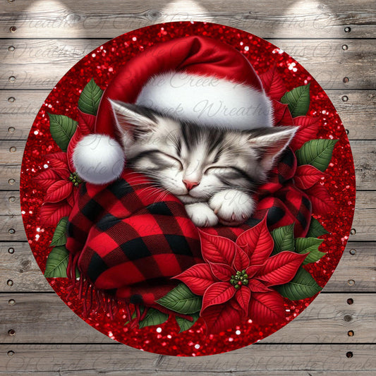 A sleeping kitten wearing a red Santa hat and wrapped in a red and black plaid blanket, surrounded by red poinsettia flowers and green leaves, all set against a wooden background with red glitter.