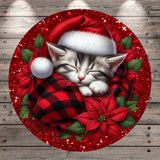 A sleeping kitten wearing a red Santa hat and wrapped in a red and black plaid blanket, surrounded by red poinsettia flowers and green leaves, all set against a wooden background with red glitter.