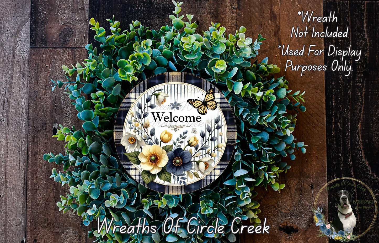 a wreath made of various green plants and flowers, with a sign that reads "Welcome" in the center. The wreath is displayed on a wooden surface, and there is a small dog in the bottom right corner of the image.