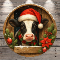 A black and white cow wearing a Santa hat and scarf peeks out from behind a wooden frame adorned with Christmas decorations, including poinsettias and ornaments.