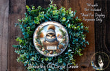 A wreath made of green foliage and pine cones, featuring a small brown squirrel wearing a plaid hat and a "sweater weather" sign.