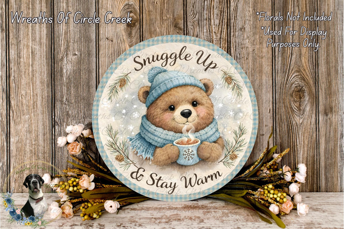 A round wooden plaque with a teddy bear wearing a blue scarf and hat, holding a cup of hot chocolate, surrounded by flowers and greenery.