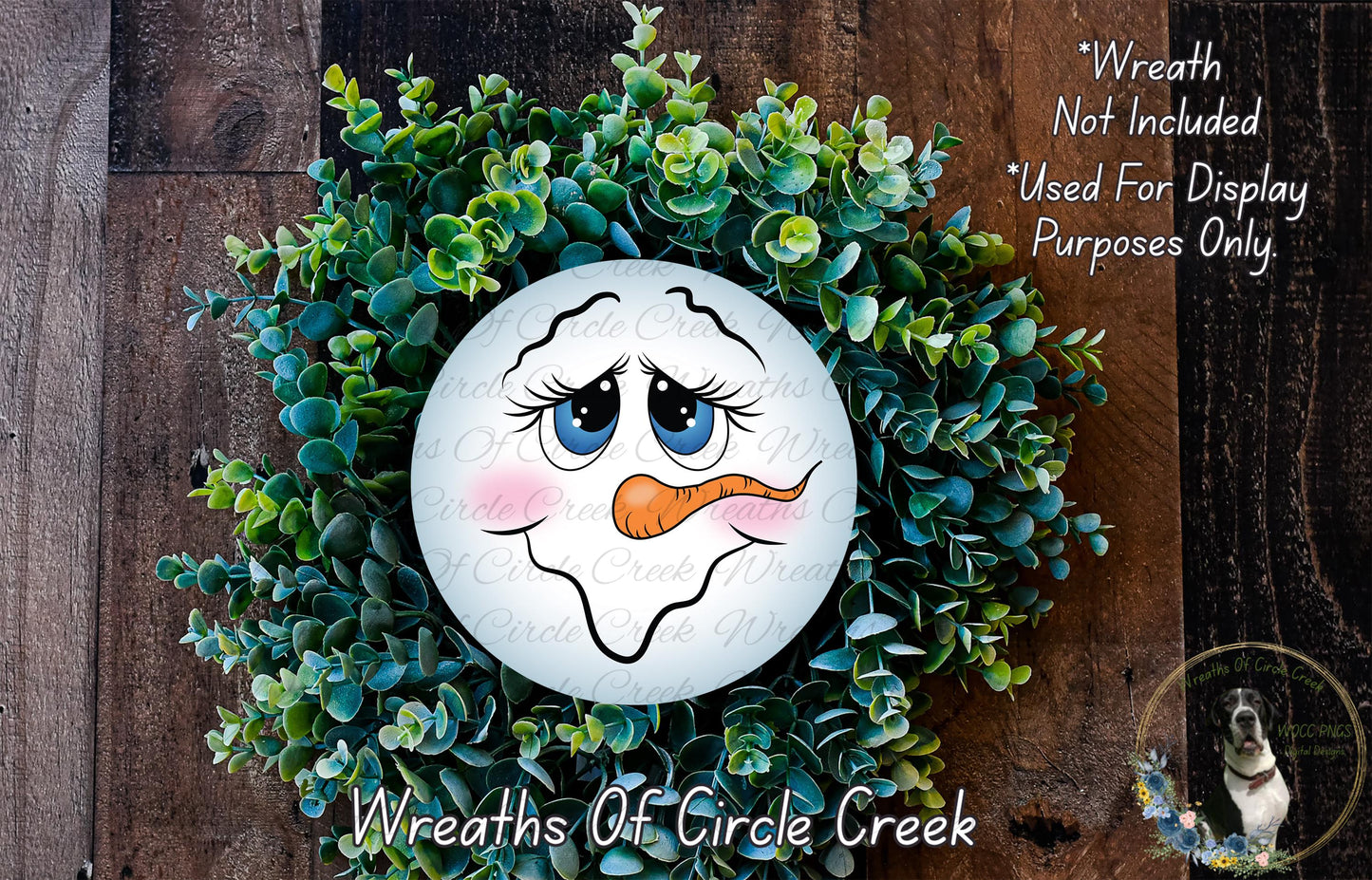 A wreath made of green plants and a white snowman face with a carrot nose, placed on a wooden surface.