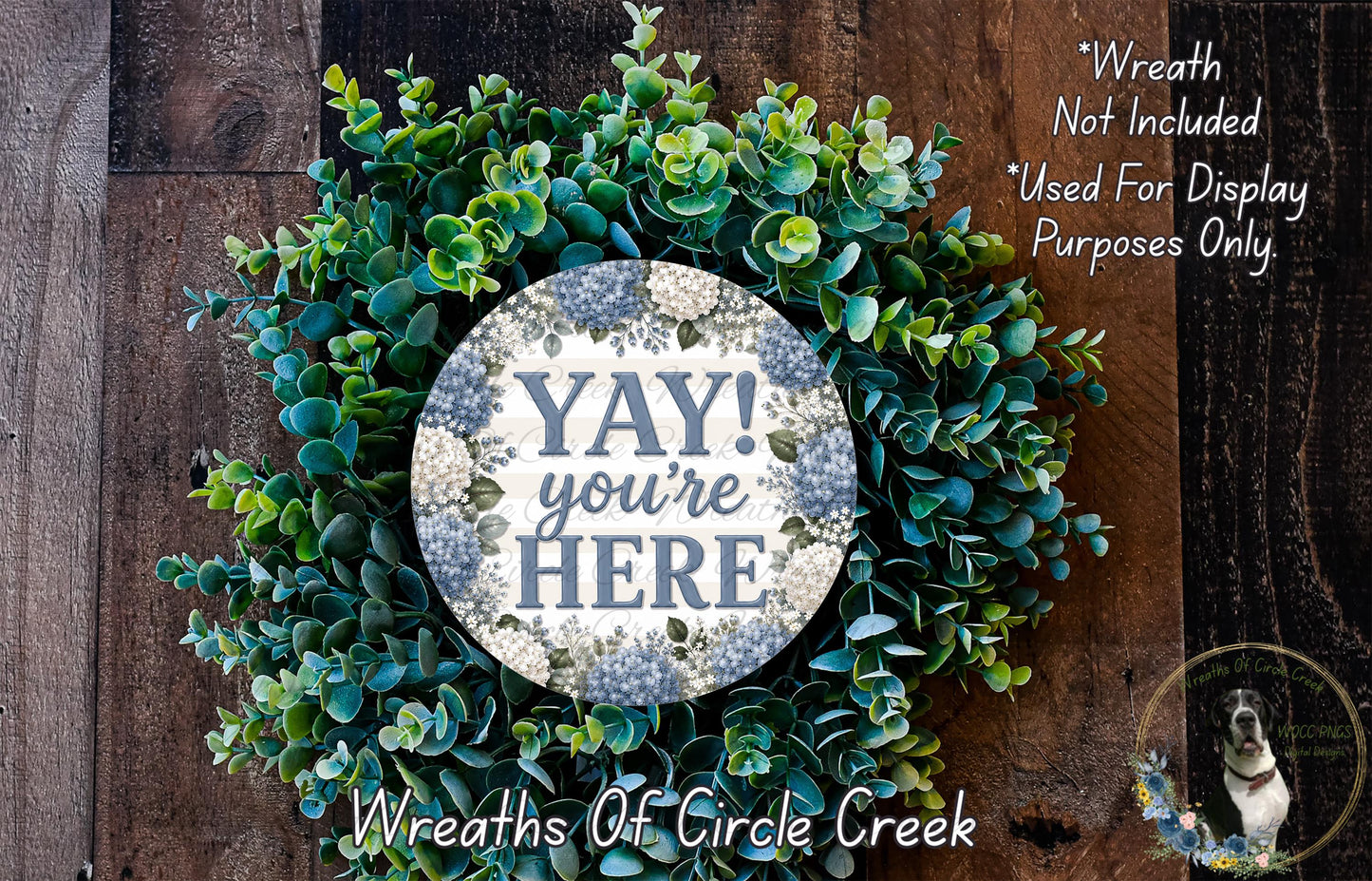 a wreath made of various green plants, including succulents and ferns, with a large circular sign that says "YAY! you're here". The sign is surrounded by the wreath, which is displayed on a wooden surface.