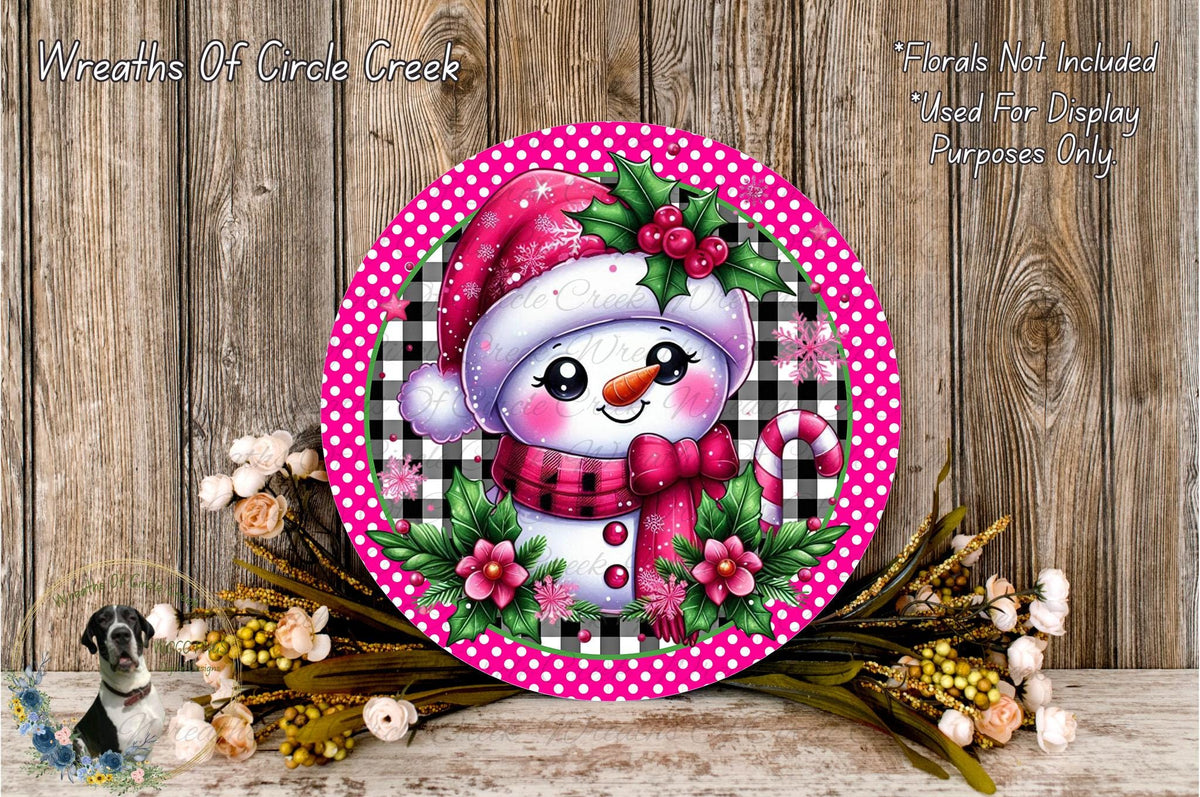 A round, decorative wreath with a snowman figure wearing a red hat and scarf, surrounded by pink and black polka dots, flowers, and a wooden background.